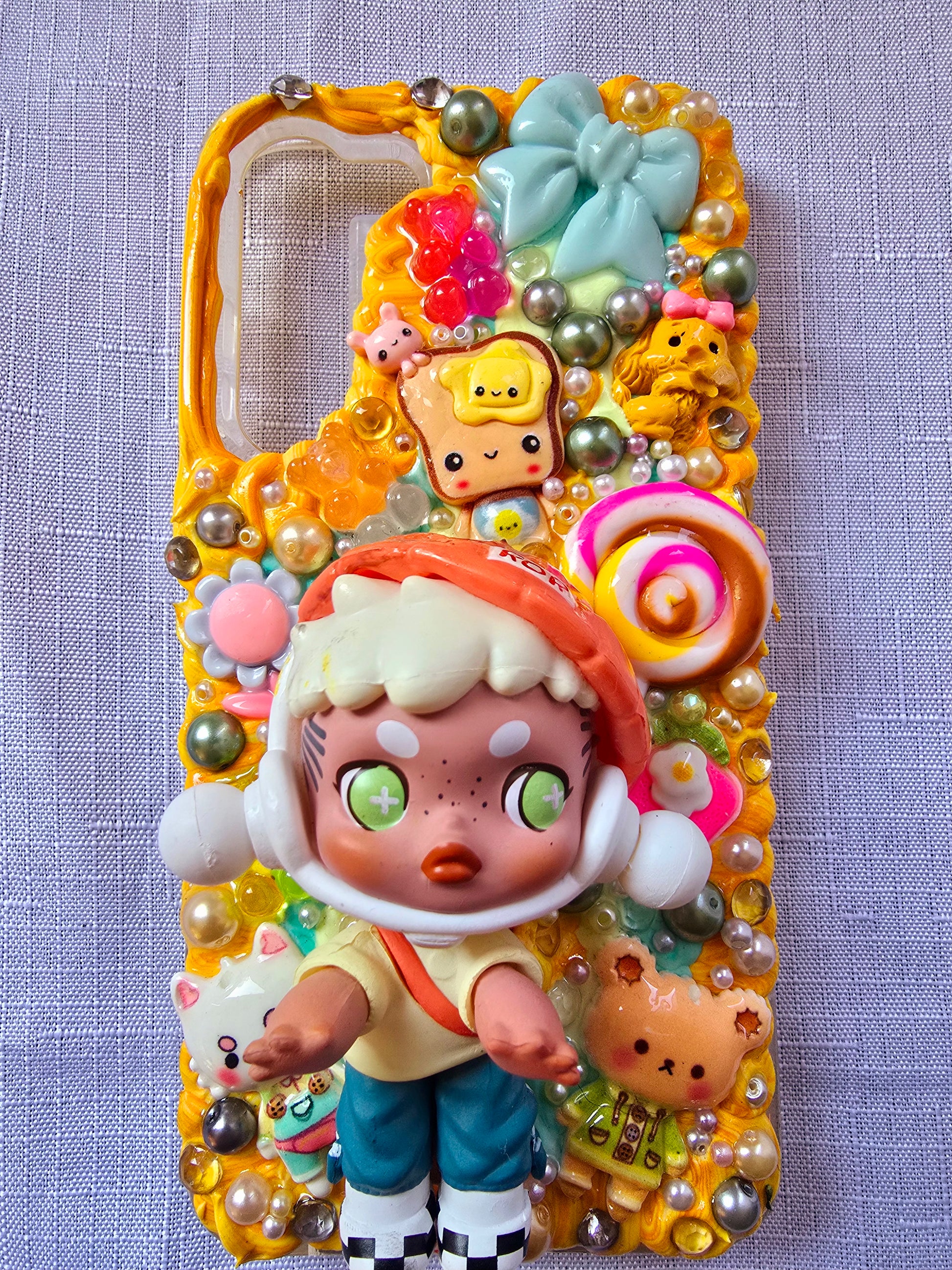 Handmade Samsung S23 Plus phone case with colorful kawaii and toy-themed 3D decorations, featuring a doll figure, pearls, cartoon charms, and candy accents on a yellow base.
