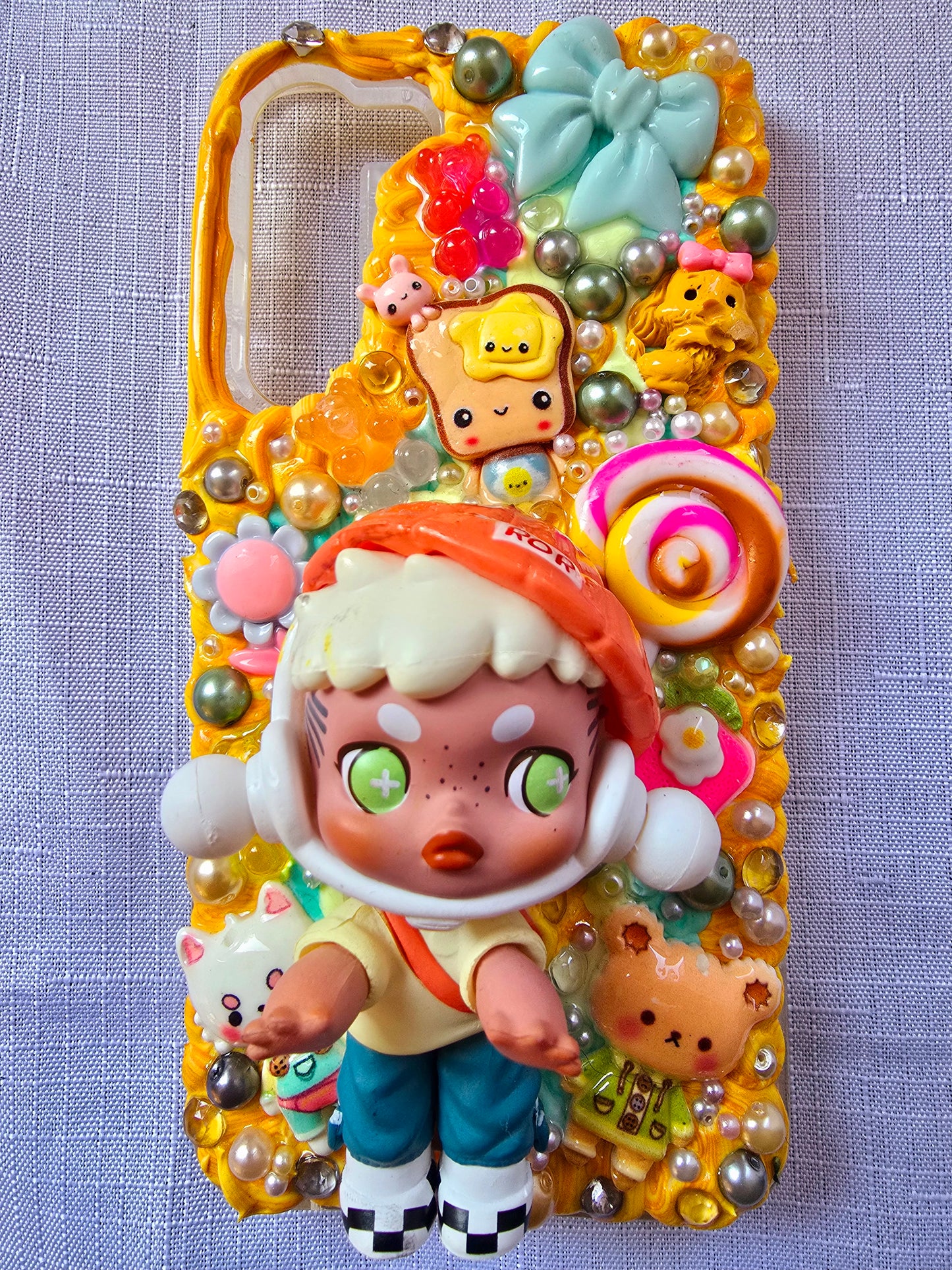 Handmade Samsung S23 Plus phone case with colorful kawaii and toy-themed 3D decorations, featuring a doll figure, pearls, cartoon charms, and candy accents on a yellow base.