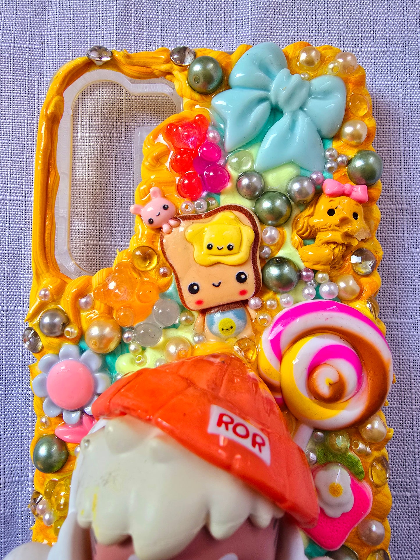 Handmade Samsung S23 Plus phone case with colorful kawaii and toy-themed 3D decorations, featuring a doll figure, pearls, cartoon charms, and candy accents on a yellow base.